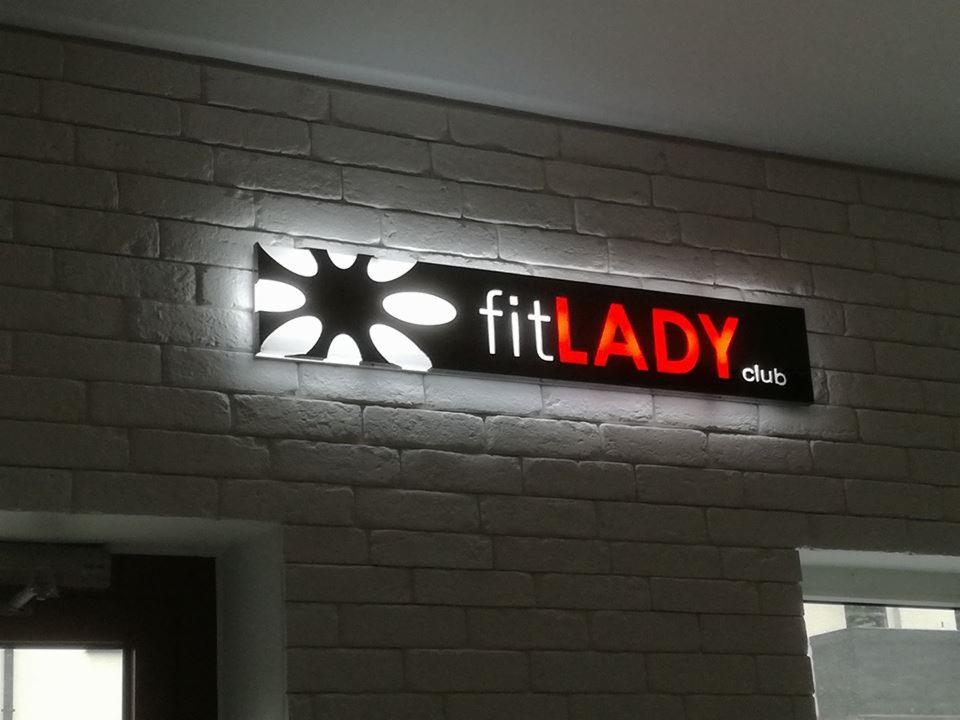 Podświetlany szyld reklamowy 'fitLADY.club' z elementem graficznym w kształcie gwiazdy umieszczony na ścianie z białej cegły wewnątrz budynku.