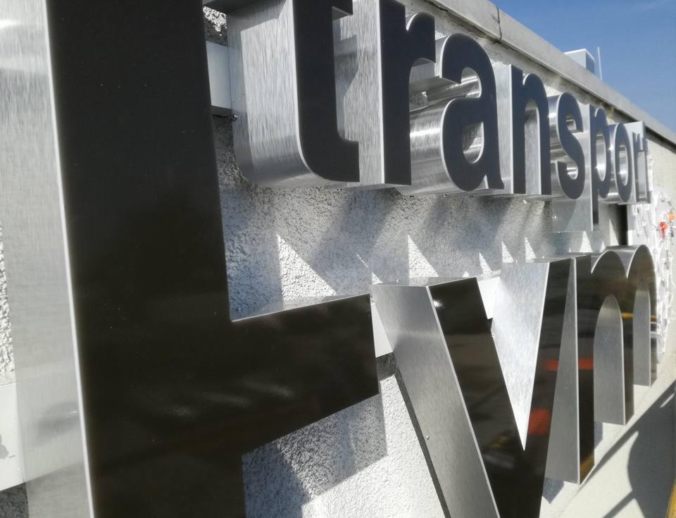 Metalowe, przestrzenne litery tworzące napis 'transport' na szorstkiej, jasnej ścianie budynku, widoczne z dołu pod kątem.