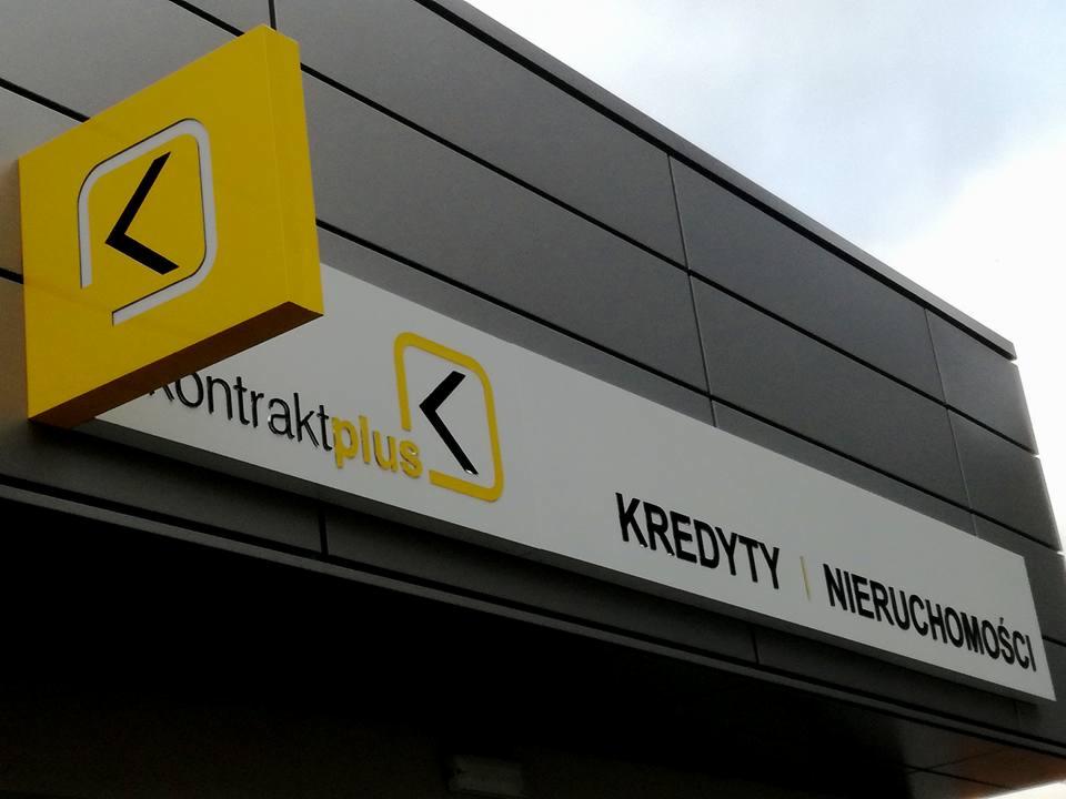 Żółto-biały szyld firmy 'Kontraktplus' z logo w kształcie strzałki w lewo, oferującej kredyty i nieruchomości, zamontowany na szarej elewacji budynku.
