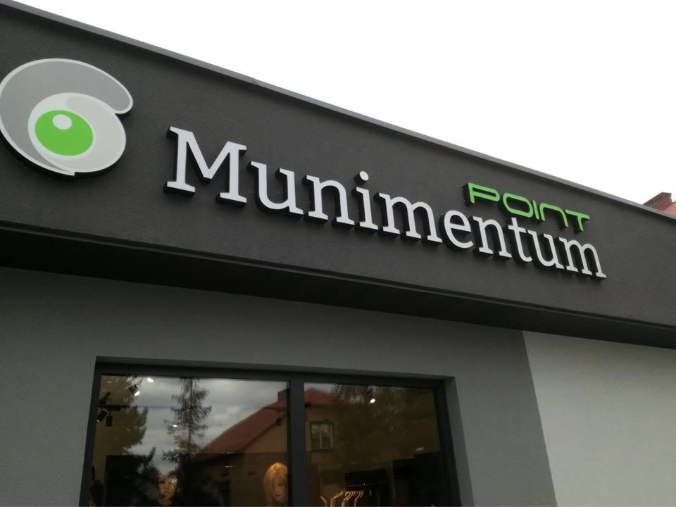 Zewnętrzna tablica z nazwą firmy Munimentum POINT z logo w kształcie oka, umieszczona nad oknem wystawowym budynku.