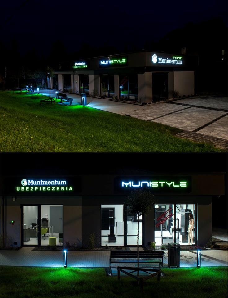 Nocny widok na budynek z podświetlanymi neonowymi szyldami firmowymi 'Munimentum Ubezpieczenia' i 'Munistyle', oświetlony zielonymi lampami, z ławkami na trawniku.