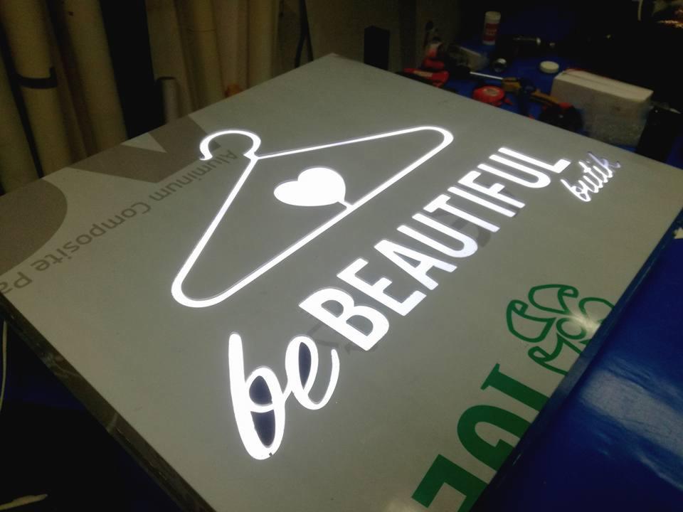 Świecący szyld reklamowy 'be BEAUTIFUL butik' z wizerunkiem wieszaka z sercem, wykonany z kompozytu aluminiowego, widoczny pod kątem na niebieskim tle, w tle narzędzia.