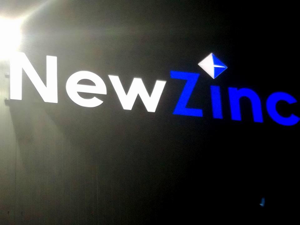 Podświetlany szyld firmy NewZinc z logo w kształcie rombu, wykonany z białego i niebieskiego akrylu, widoczny w nocy.