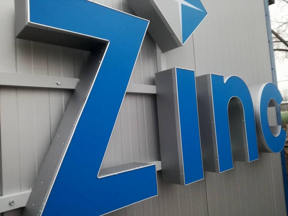 Zewnętrzny szyld reklamowy firmy 'Zinc' z niebieskimi literami 3D i szarą obudową, zamontowany na szarej ścianie z widocznymi elementami mocującymi.