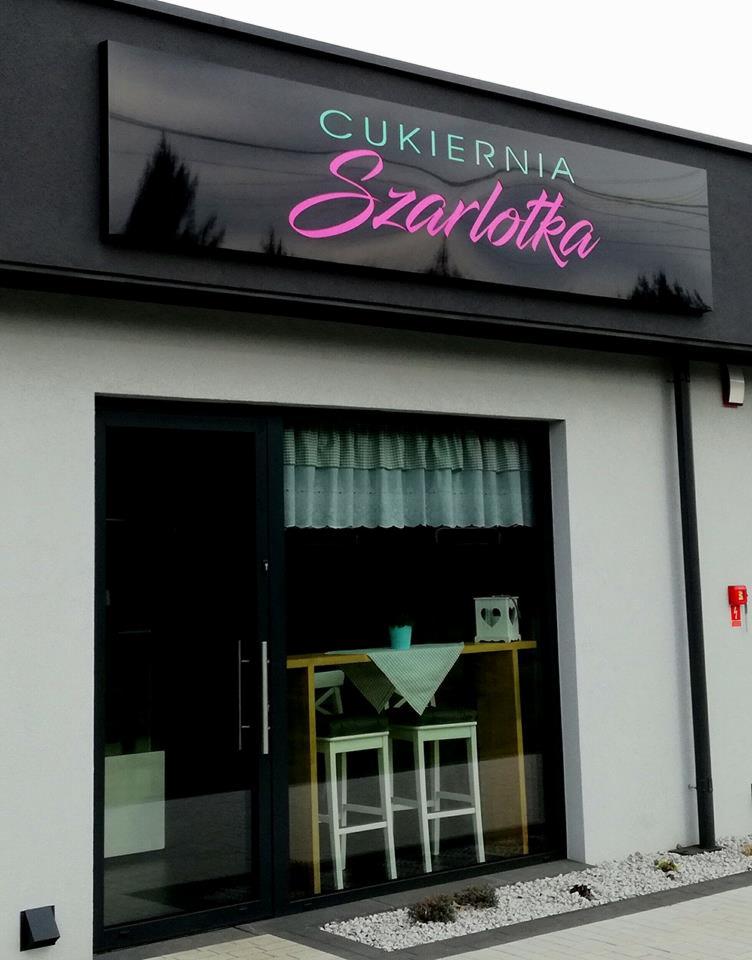 Elewacja cukierni 'Szarlotka' w Pszczynie z czarnym szyldem z różowym napisem i widocznym wnętrzem przez szklane drzwi.