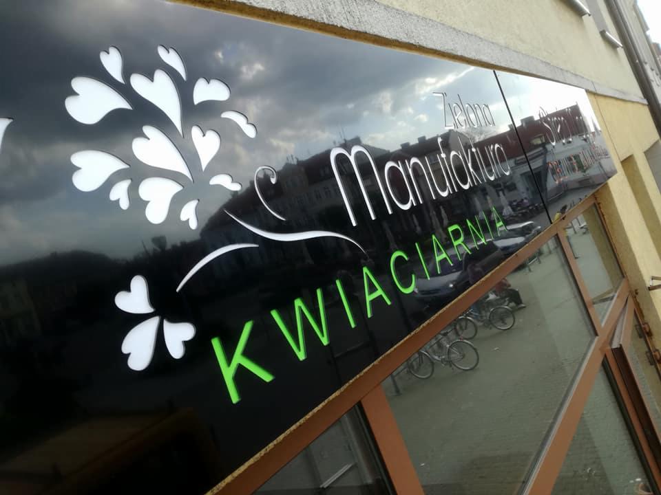 Zewnętrzny szyld kwiaciarni 'Zielona Manufaktura' z białymi elementami graficznymi w kształcie serc i zielonym napisem 'KWIACIARNIA', odbijający ulicę w błyszczącej powierzchni.