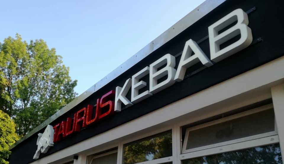 Zewnętrzny szyld reklamowy 'TAURUS KEBAB' z podświetlanymi literami, umieszczony na czarnym tle budynku z białymi obramowaniami okien, widok z dołu, z drzewami w tle.
