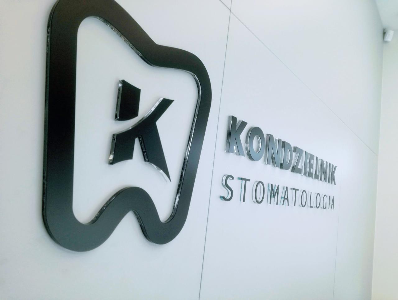 Logo gabinetu stomatologicznego: czarny obrys zęba z literą K w środku i srebrny napis 'KONDZIELNIK STOMATOLOGIA' na białej ścianie.