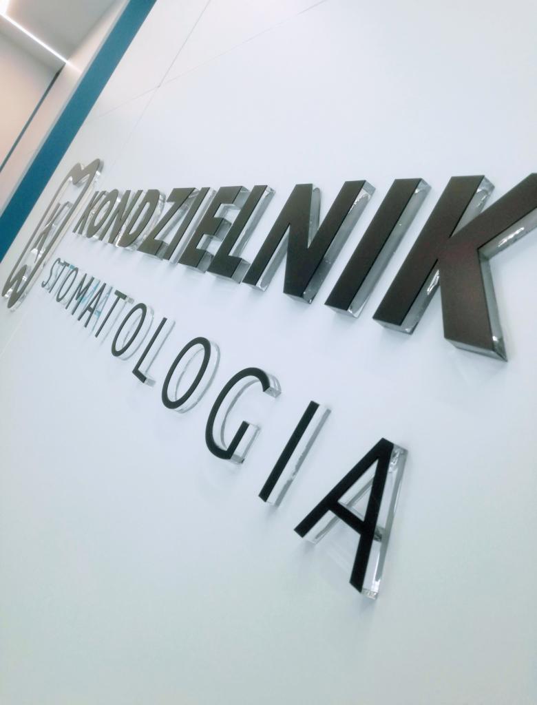 Elegancki napis 3D 'Kondzielnik Stomatologia' z czarnymi literami z przezroczystym akrylowym obramowaniem, zamontowany na białej ścianie w klinice dentystycznej, widok z dołu.