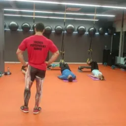 Trener personalny Dawid nadzoruje ćwiczenia na TRX w siłowni z pomarańczową podłogą, widziany od tyłu w koszulce z napisem 'TRENER PERSONALNY DAWID' i legginsach z nadrukiem mięśni.