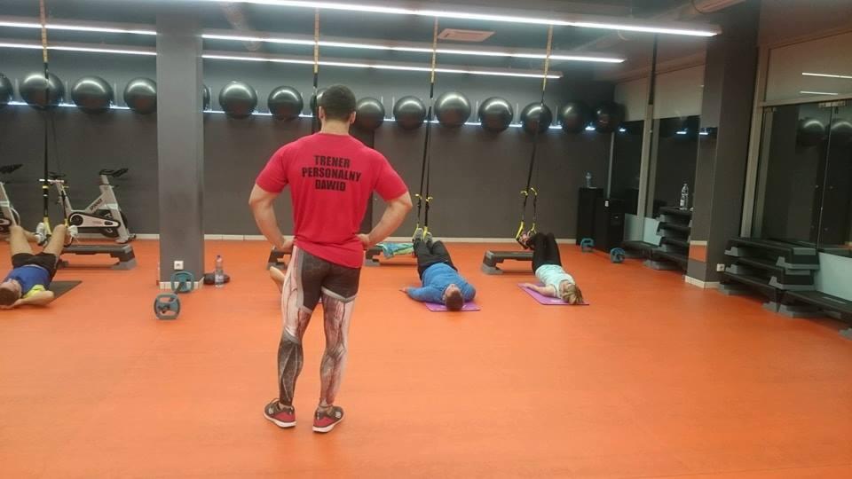 Trener personalny Dawid nadzoruje ćwiczenia na TRX w siłowni z pomarańczową podłogą, widziany od tyłu w koszulce z napisem 'TRENER PERSONALNY DAWID' i legginsach z nadrukiem mięśni.