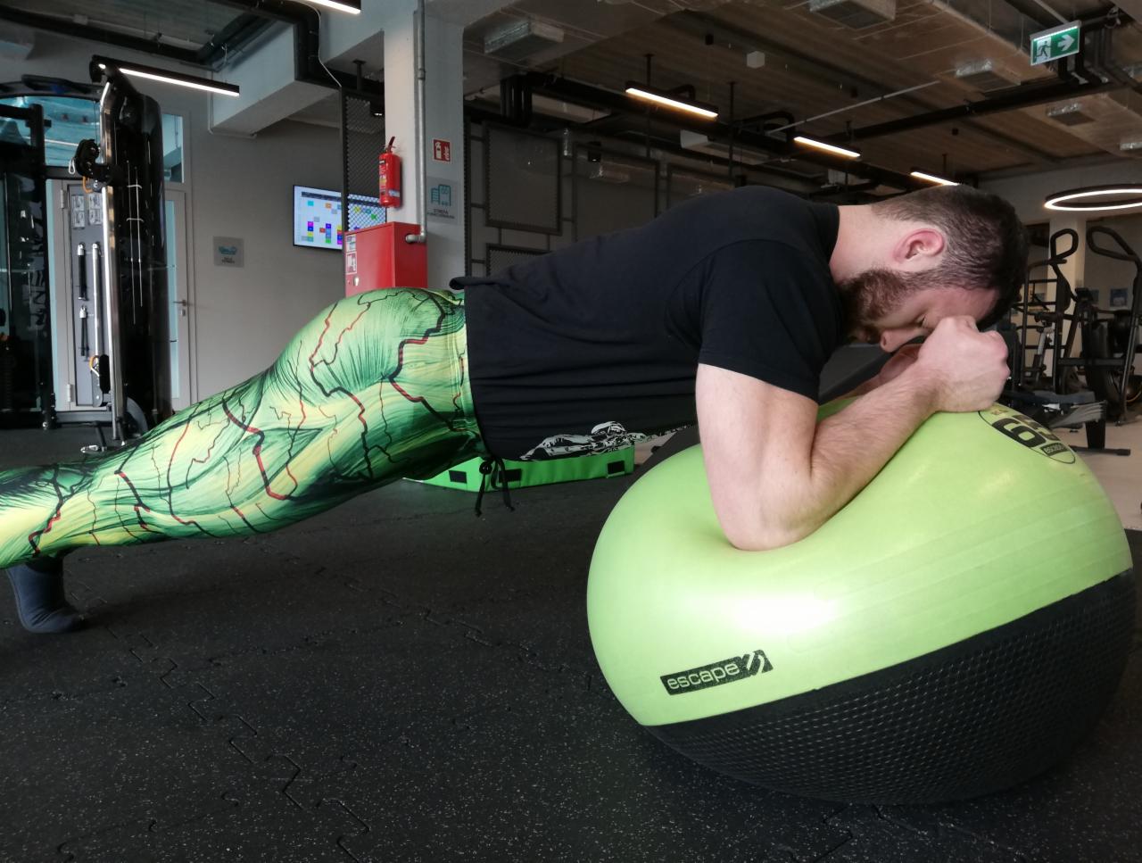 Mężczyzna w legginsach z zielonym wzorem wykonuje ćwiczenie plank na piłce do ćwiczeń w siłowni.