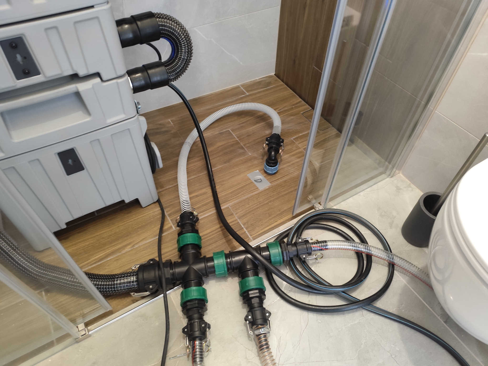 Szary osuszacz kondensacyjny podłączony do instalacji w łazience z kabiną prysznicową i toaletą. Węże i złączki z zielonymi elementami. Widok z góry.