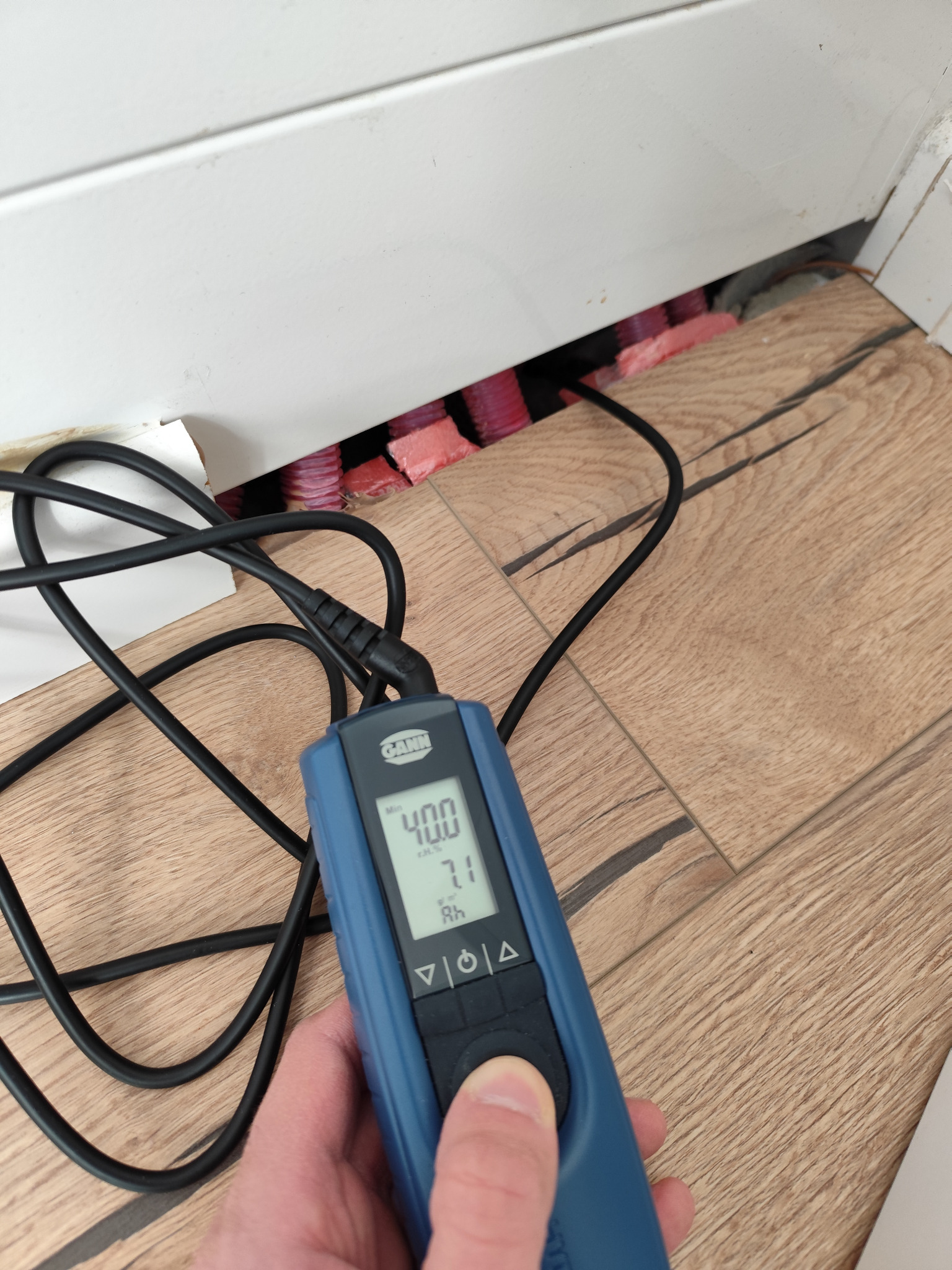 Pomiar wilgotności podłogi przy listwie przypodłogowej za pomocą wilgotnościomierza elektronicznego GANN, widoczne rury ogrzewania podłogowego.