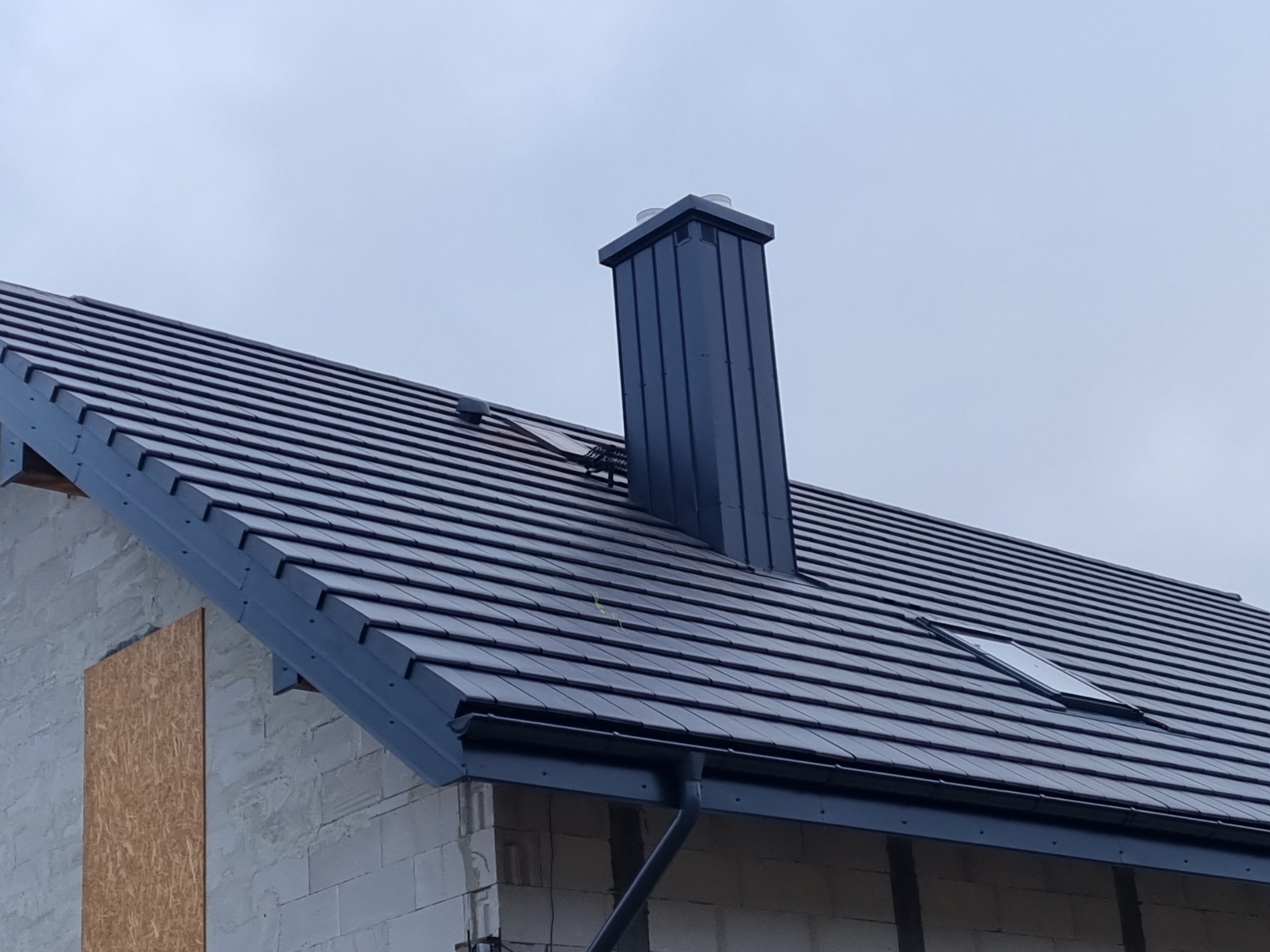 Ciemna dachówka ceramiczna na skośnym dachu domu jednorodzinnego z oknem dachowym i kominem w kolorze grafitowym, widok z dołu na tle pochmurnego nieba.