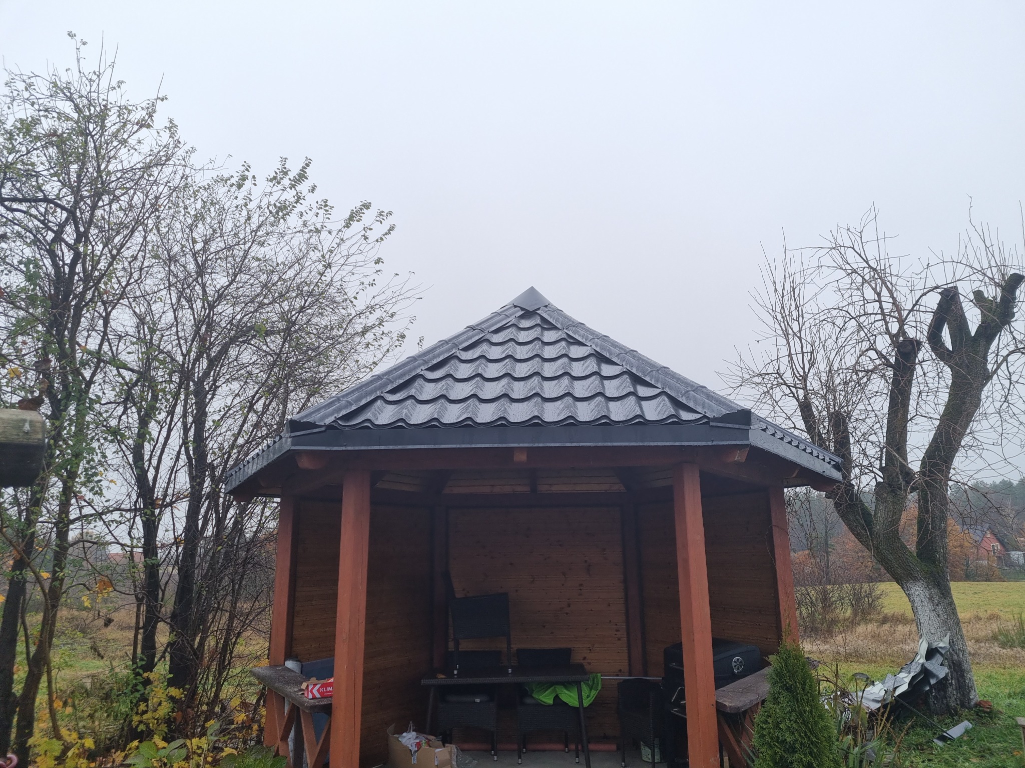 Zadaszona altana z ciemną, metalową dachówką w ogrodzie. Widoczne drzewa bez liści i ponure, szare niebo. Wnętrze altany częściowo wyposażone.