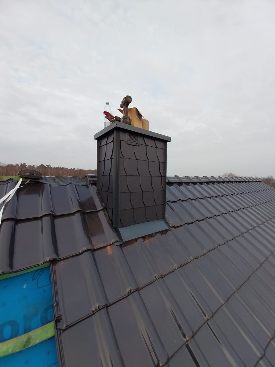 Nowy, ciemny dach z ceramicznej dachówki, z widocznym kominem wykończonym w podobnym stylu. Na kominie narzędzia i butelka. Tło: pochmurne niebo i drzewa.