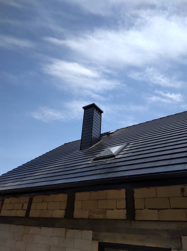 Nowoczesny dach z czarnej dachówki ceramicznej, komin z blachy w kolorze dachu i okno dachowe na tle błękitnego nieba.