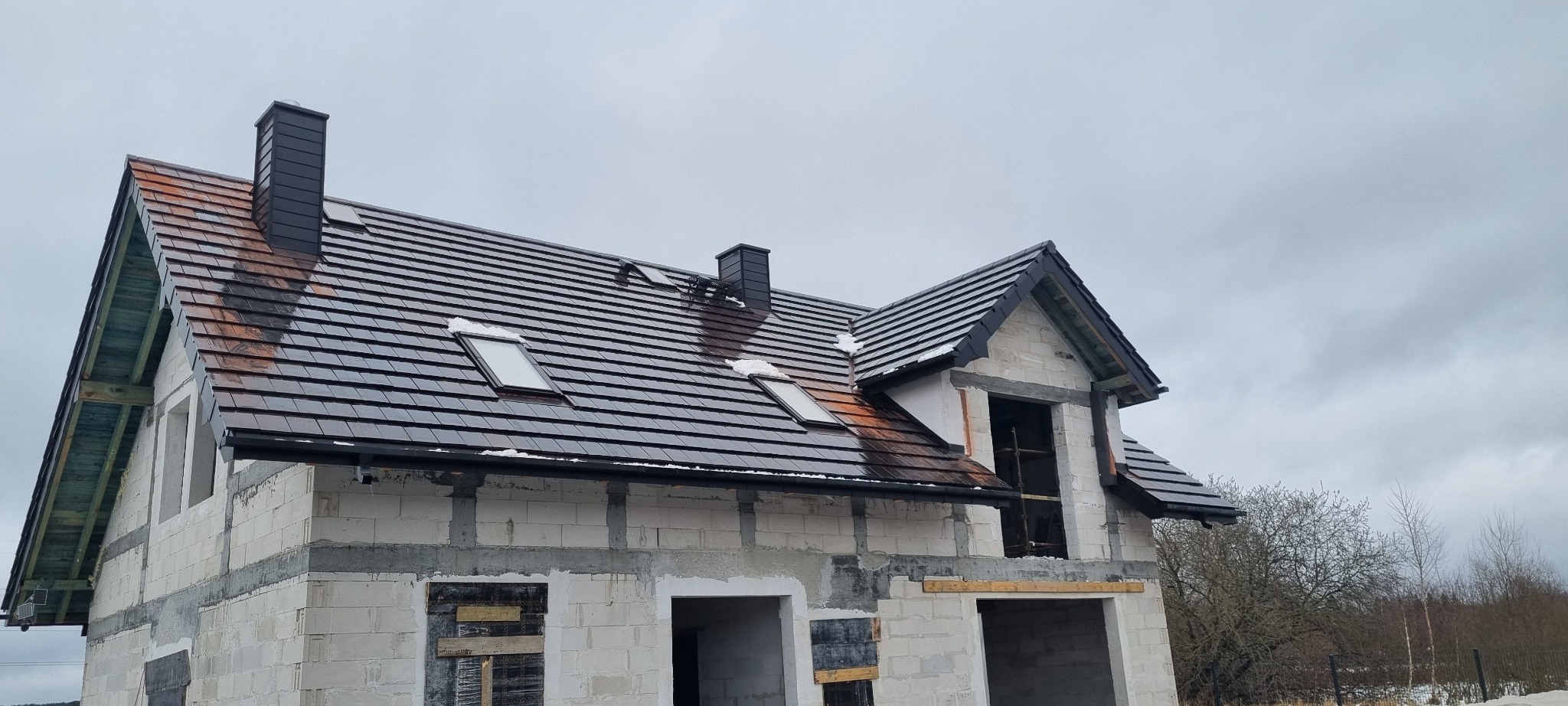Niedokończony dom z ciemną dachówką, widoczne okna dachowe i kominy. Budynek w stanie surowym, ściany z białego pustaka.