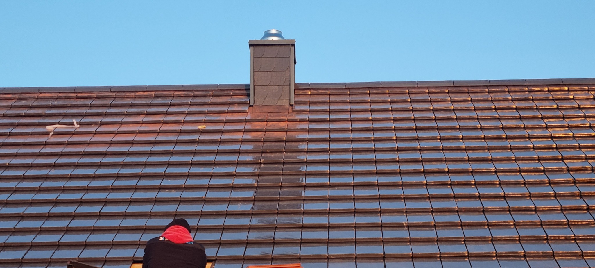 Błyszczący dach z brązowej dachówki ceramicznej w regularnym wzorze, z kominem i fragmentem osoby w dolnej części kadru na tle błękitnego nieba.