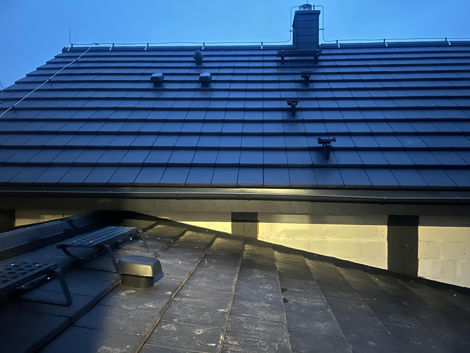 Nowy dach z czarnej dachówki ceramicznej, widok z dołu. Zamontowane stopnie kominiarskie i odpowietrzniki. Ujęcie wykonane o zmierzchu.