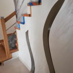 Ściana z dekoracyjną, falistą wnęką, obok schody zabezpieczone folią i taśmą malarską, przygotowane do prac wykończeniowych.