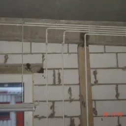 Surowe wnętrze z widocznymi, poprowadzonymi po ścianie i suficie rurami instalacji elektrycznej, ściany z białego bloczka betonowego, okno z widokiem na zewnątrz.