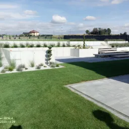 Nowoczesny ogród z betonowymi rabatami, ozdobnymi trawami i krzewami na żwirowym podłożu, z widocznym trawnikiem i ścieżką z szarej kostki brukowej, w tle dom jednorodzinny i błękitne niebo...