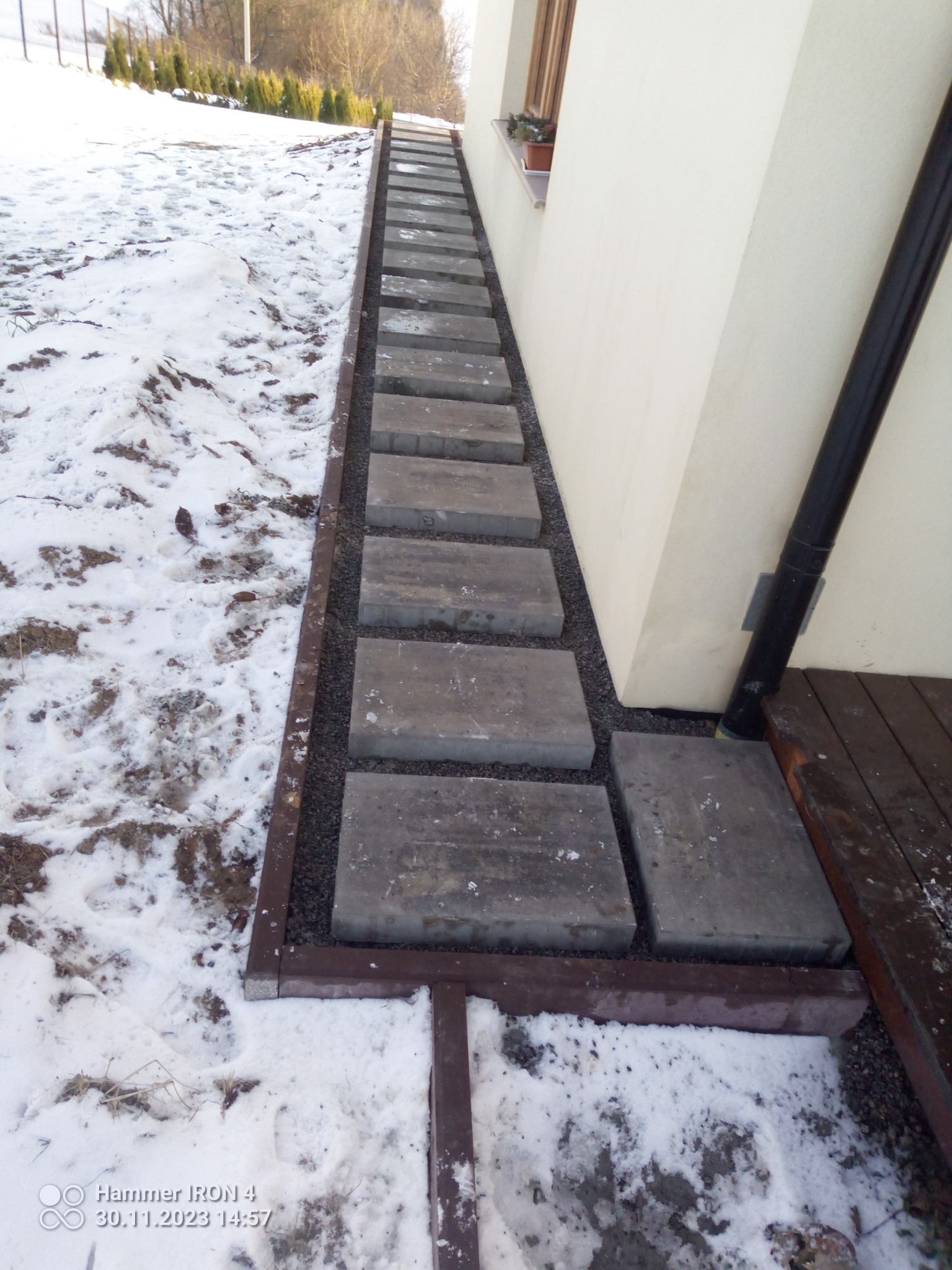 Ułożona ścieżka z betonowych płyt w ogrodzie pokrytym śniegiem, prowadząca wzdłuż ściany budynku z doniczką w oknie.