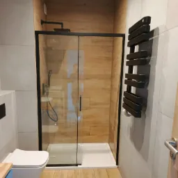 Nowoczesna kabina prysznicowa z czarnymi ramami, drewnopodobne płytki na ścianie, grzejnik drabinkowy i wisząca toaleta. Minimalistyczny design łazienki.