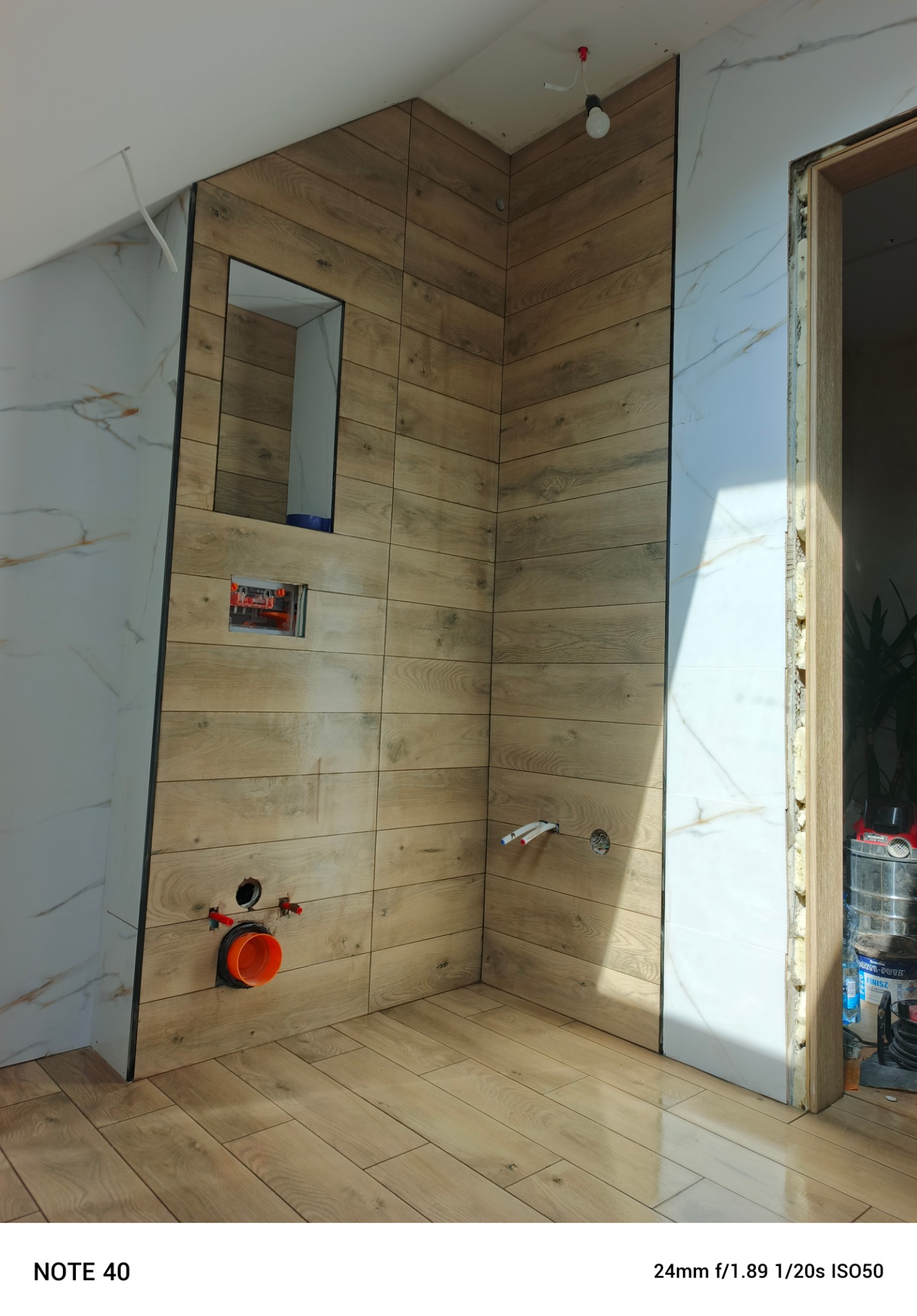Wykończenie łazienki w trakcie: drewnopodobne płytki na ścianie z wnęką i instalacją, białe płytki z żyłkami. Widoczne narzędzia i materiały budowlane.