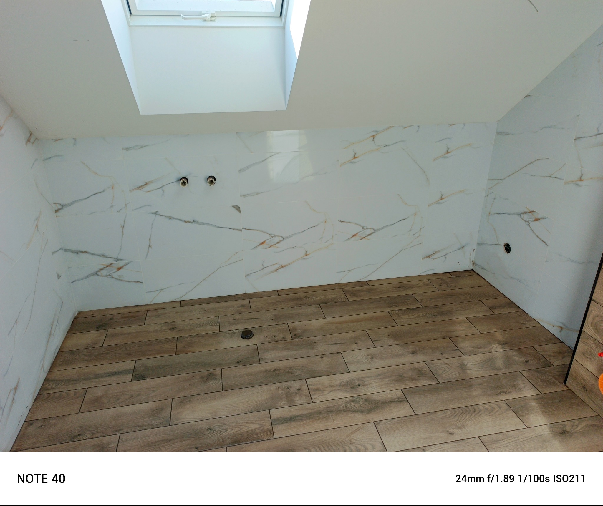 Wykończona łazienka z drewnopodobną podłogą i marmurowymi płytkami ściennymi, widok na okno dachowe i instalacje wodne. Ukończone prace wykończeniowe.