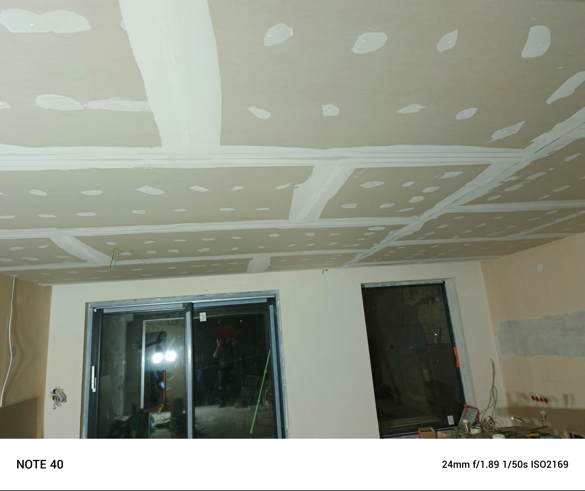 Sufit z płyt gipsowo-kartonowych w trakcie szpachlowania i wygładzania, widoczne łączenia płyt. Ściany w surowym stanie, okna przesuwne.