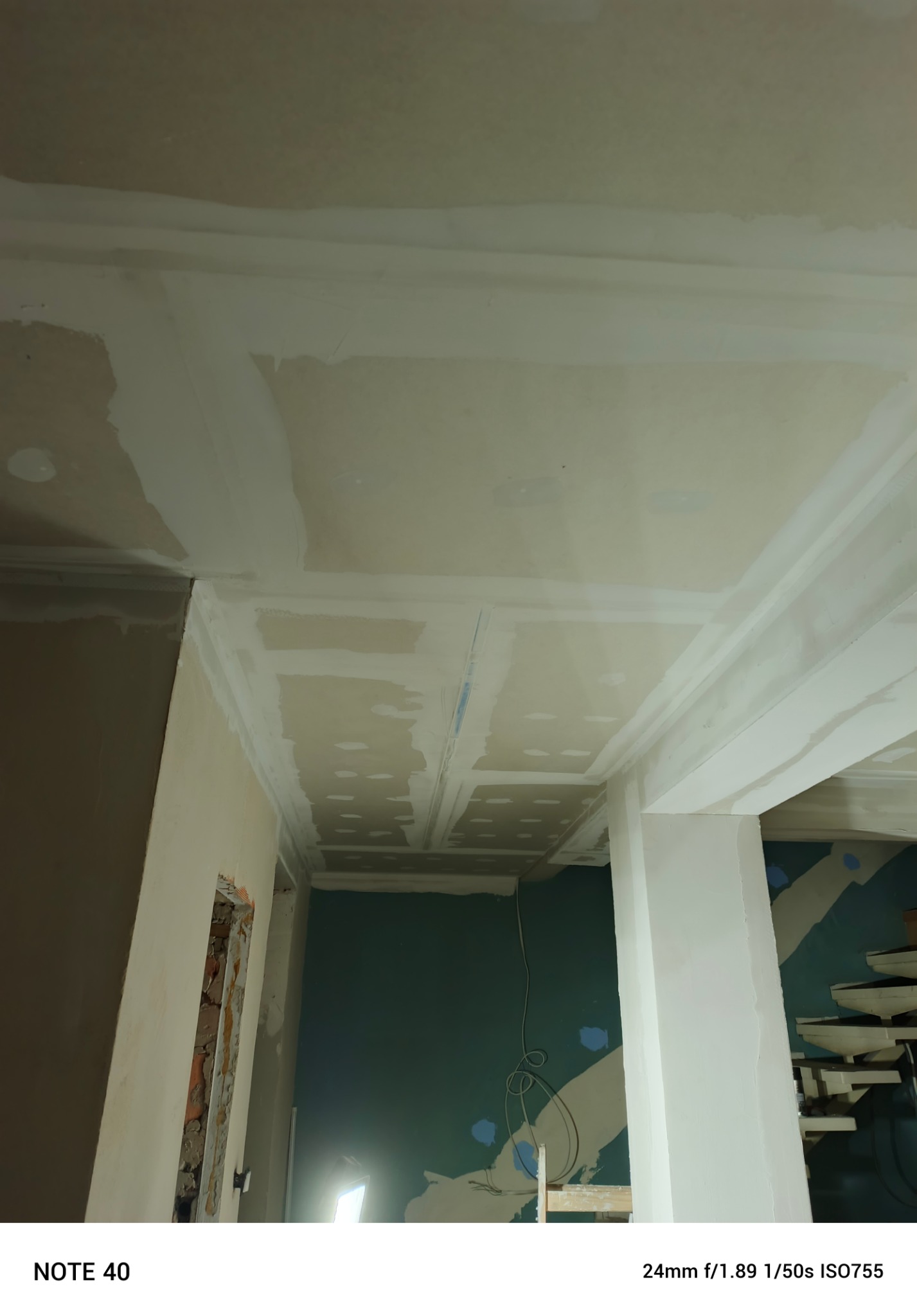 Sufit po szpachlowaniu z widocznymi płytami gipsowymi i listwami dekoracyjnymi. Ściany w trakcie przygotowania do malowania. Widoczne niedoskonałości i surowy stan.