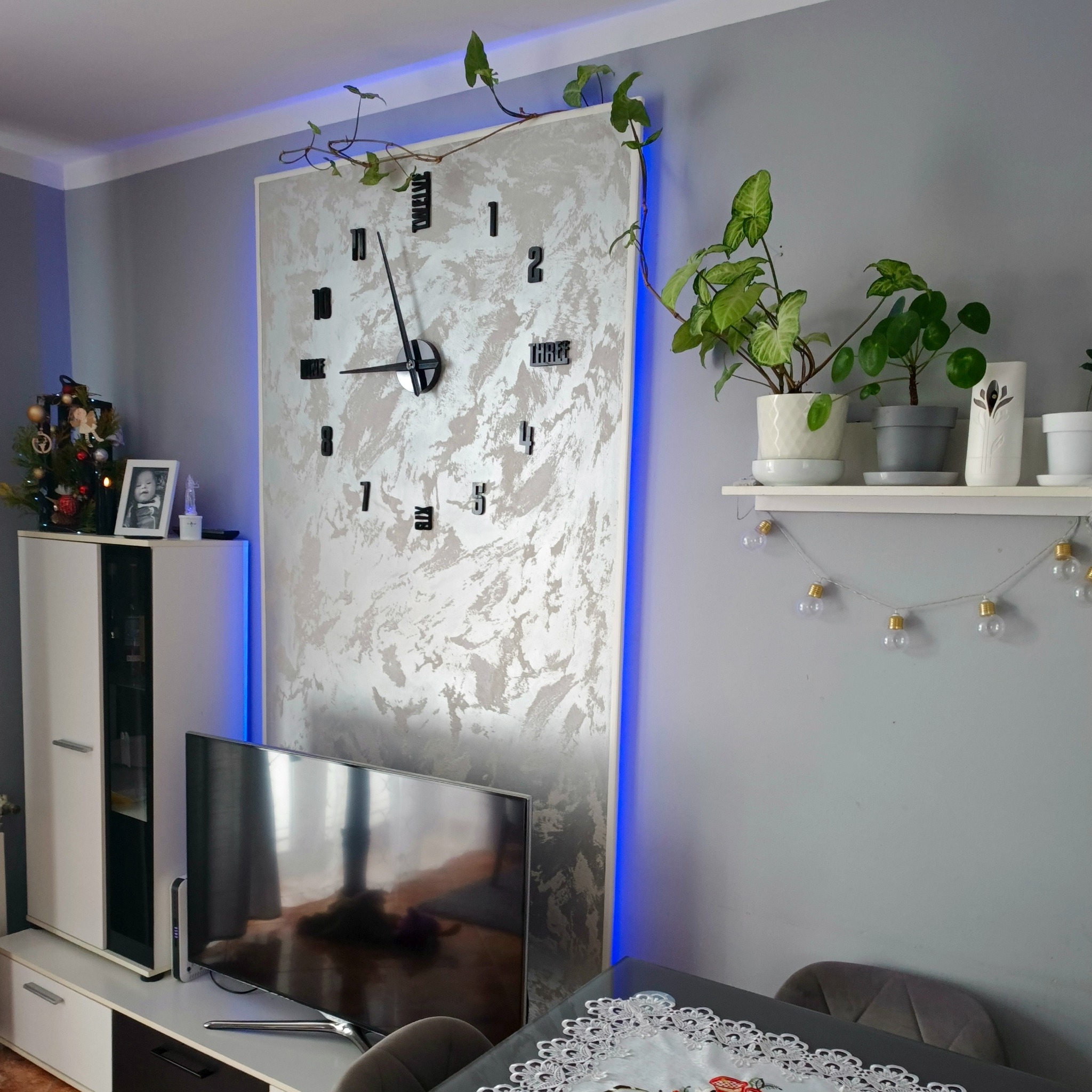 Ściana akcentowa z dekoracyjnym zegarem i podświetleniem LED w nowoczesnym salonie. Półka z roślinami, telewizor i szafka w tle.