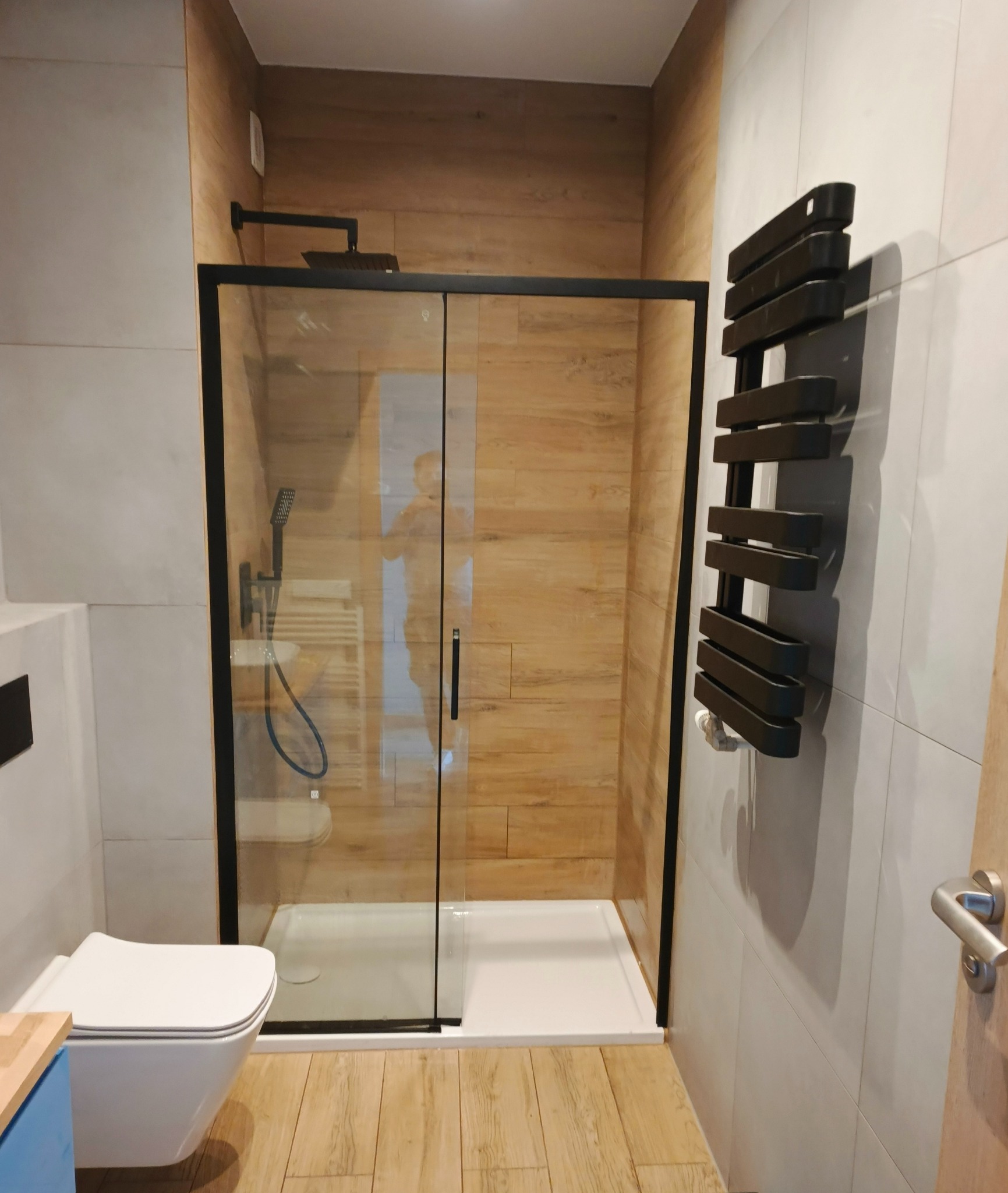 Nowoczesna kabina prysznicowa z czarnymi ramami, drewnopodobne płytki na ścianie, grzejnik drabinkowy i wisząca toaleta. Minimalistyczny design łazienki.