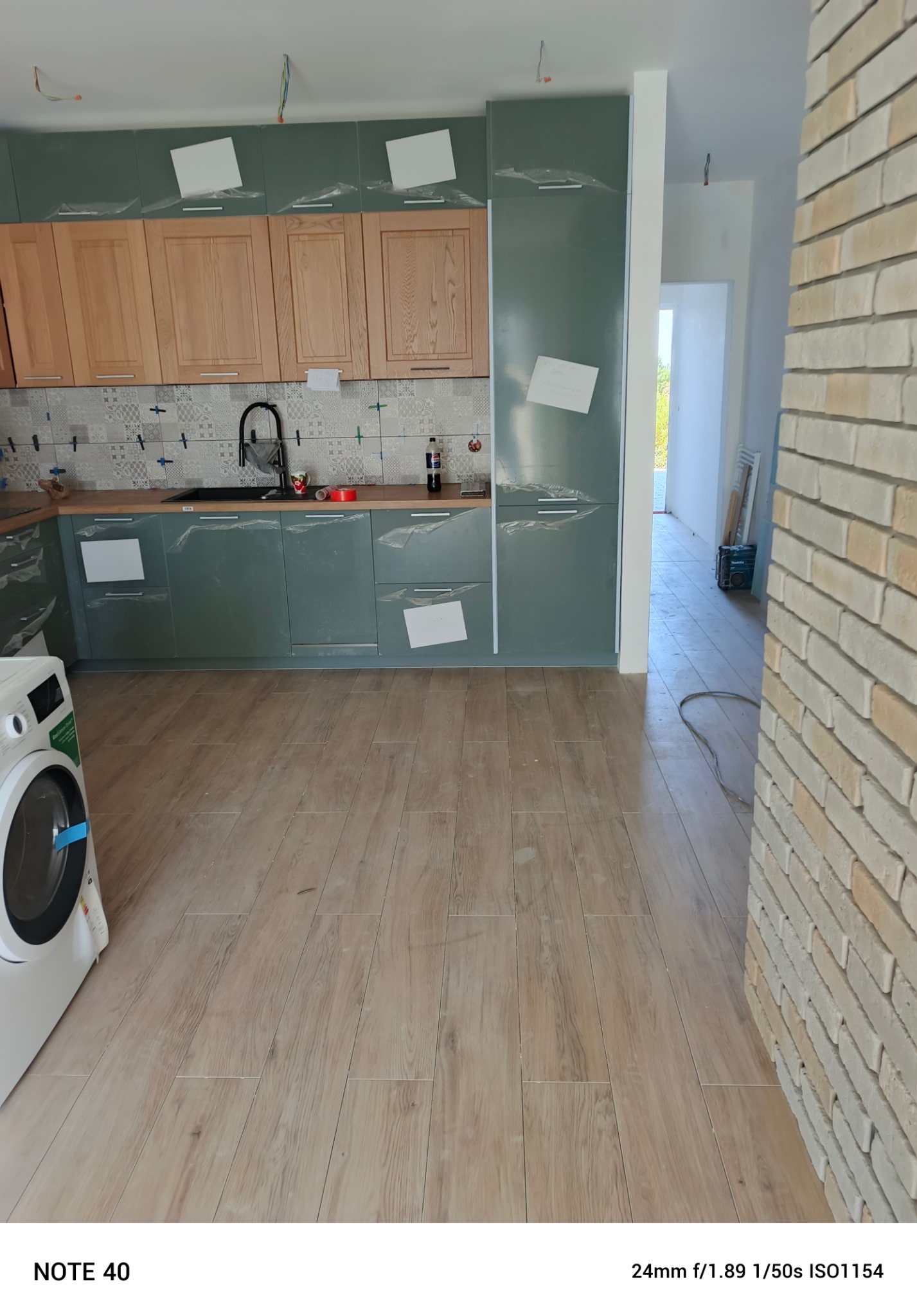 Kuchnia w trakcie montażu: zielone szafki z drewnianymi frontami, zabezpieczone folią, płytki na ścianie, pralka i drewnopodobna podłoga.