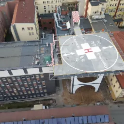 Widok z góry na nowo wybudowane lądowisko dla helikopterów na dachu budynku szpitala Copernicus, z oznaczeniami i strzałkami, otoczone budynkami z czerwonej cegły i dachem z szachownicą.