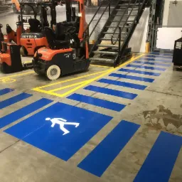 Oznakowanie hali produkcyjnej z wyznaczonym przejściem dla pieszych w kolorze niebieskim i żółtym, widoczne wózki widłowe i schody metalowe.