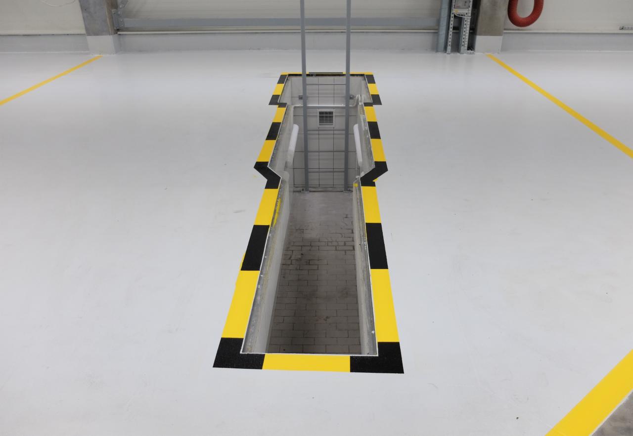 Jasnoszara posadzka przemysłowa z wyznaczonymi żółtymi liniami i obrzeżem wokół kanału rewizyjnego z czarno-żółtymi pasami ostrzegawczymi.