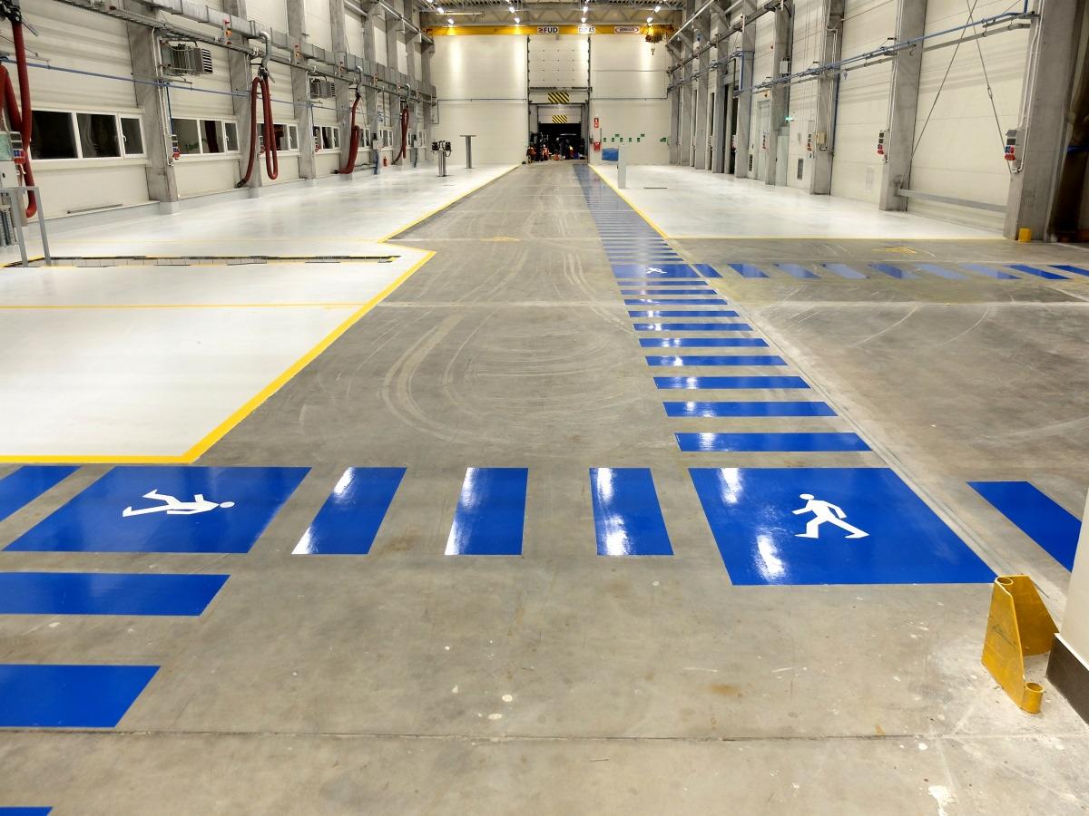Wnętrze hali przemysłowej z szarą posadzką, wyznaczona niebieska ścieżka dla pieszych z symbolami postaci, żółte linie oddzielające strefy, widoczne instalacje pod sufitem.