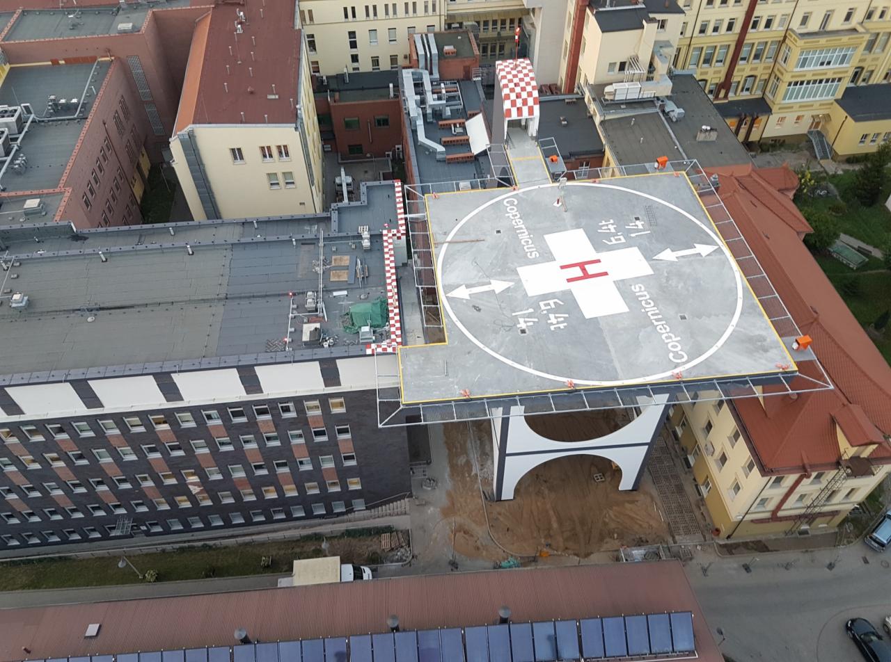 Widok z góry na nowo wybudowane lądowisko dla helikopterów na dachu budynku szpitala Copernicus, z oznaczeniami i strzałkami, otoczone budynkami z czerwonej cegły i dachem z szachownicą.