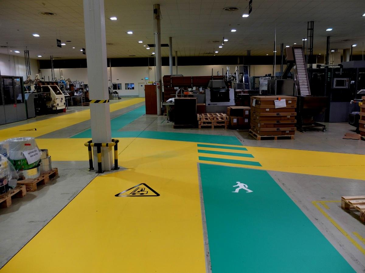 Wnętrze hali produkcyjnej z żółto-zieloną posadzką, wyznaczonymi ciągami komunikacyjnymi i znakami ostrzegawczymi na podłodze.