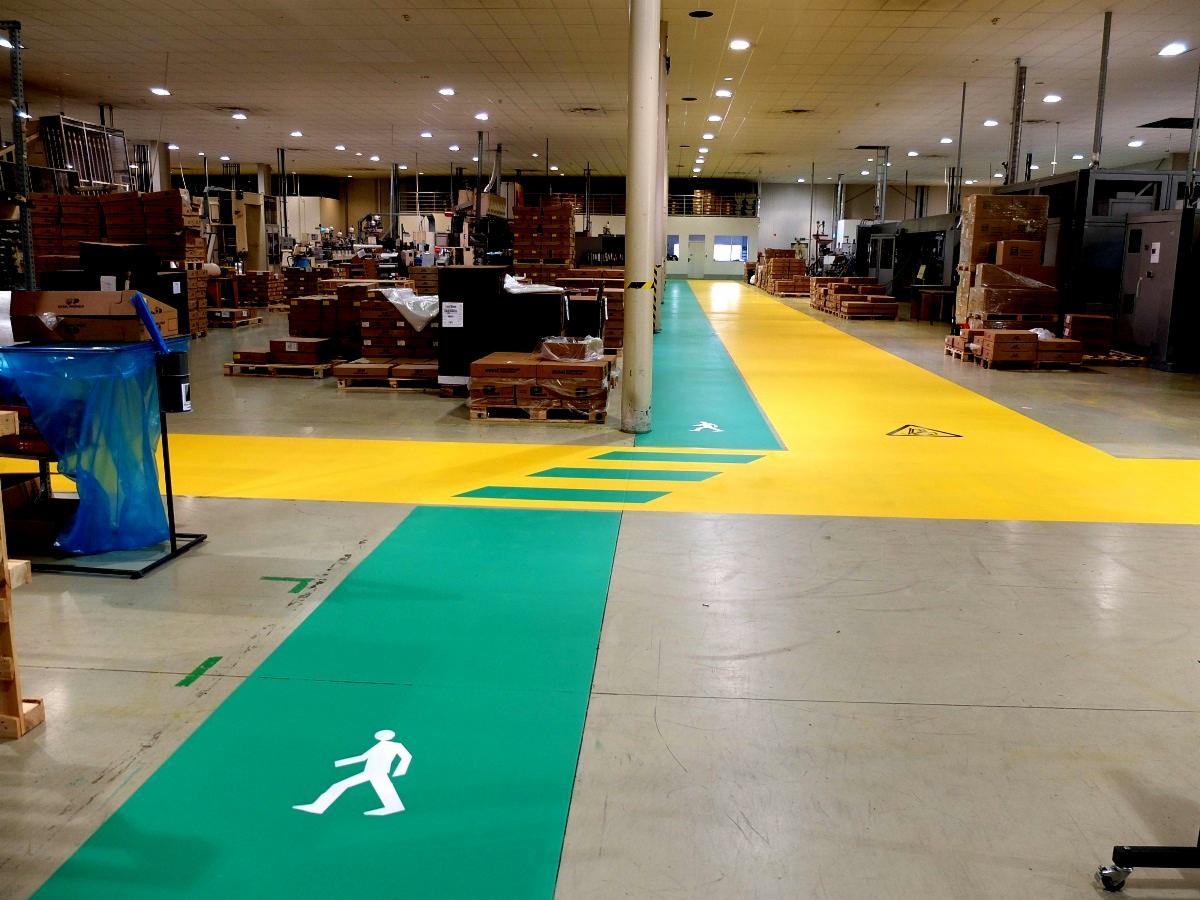 Oznakowanie poziome w hali produkcyjnej, z zielonymi ścieżkami dla pieszych i żółtymi strefami, kolumna w tle, skrzynie i maszyny w oddali.