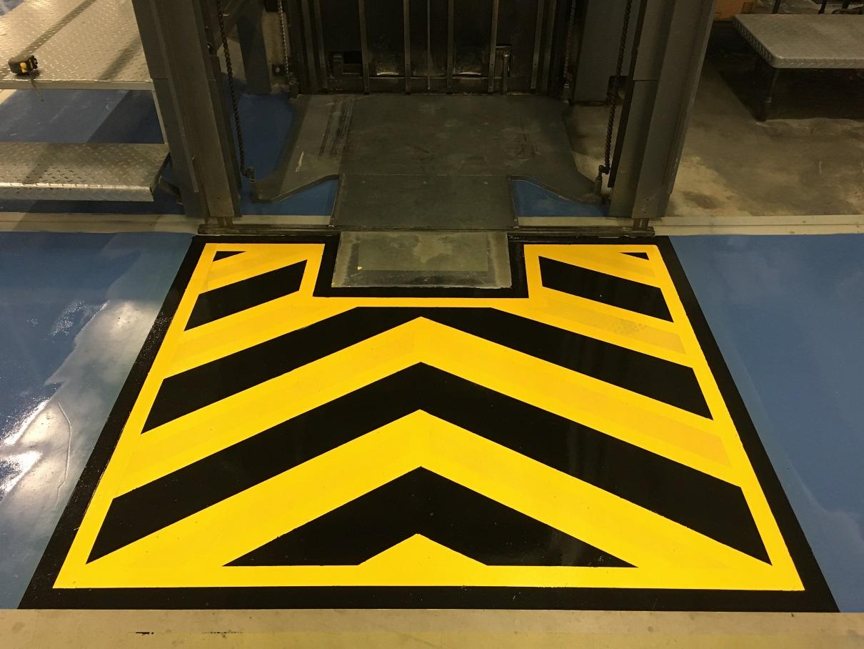 Oznakowanie strefy niebezpiecznej żółto-czarnymi pasami na posadzce przemysłowej przed bramą wjazdową.