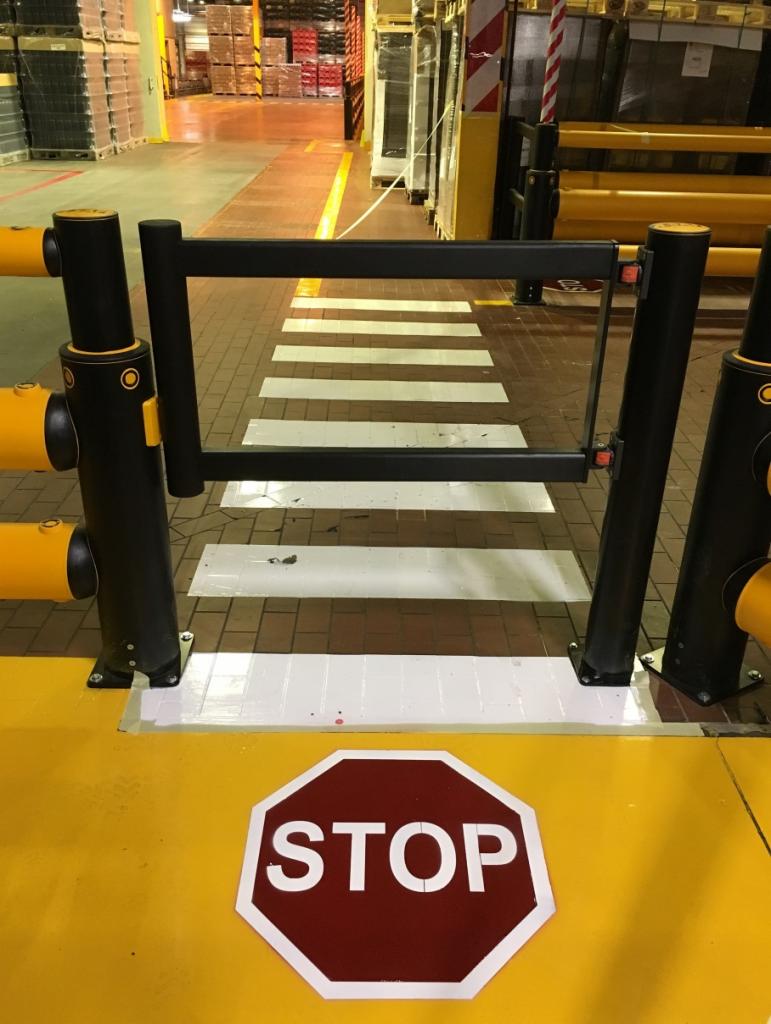 Oznakowanie poziome w hali produkcyjnej: przejście dla pieszych z białymi pasami, barierki ochronne z żółtymi elementami i znak STOP na żółtej posadzce.