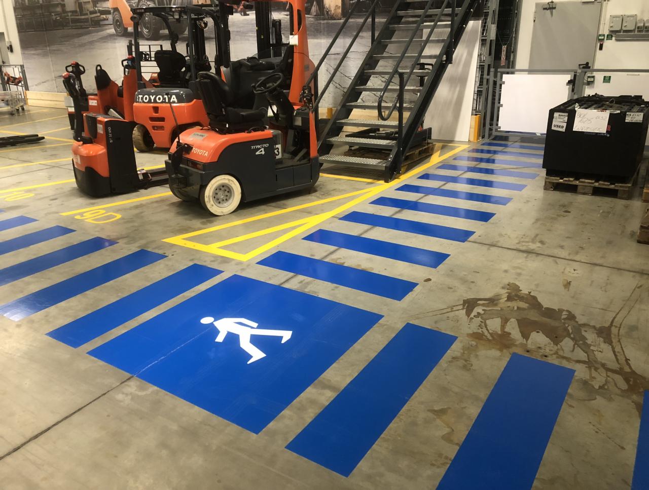 Oznakowanie hali produkcyjnej z wyznaczonym przejściem dla pieszych w kolorze niebieskim i żółtym, widoczne wózki widłowe i schody metalowe.