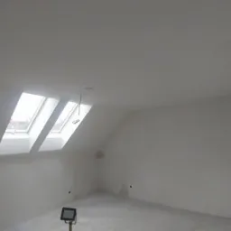 Poddasze po szpachlowaniu i przygotowaniu do malowania, widoczne dwa okna dachowe, surowe ściany, lampa budowlana na statywie oraz wystające kable.
