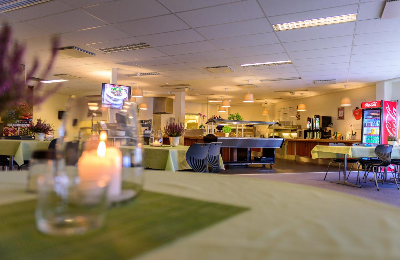 Wnętrze restauracji lub stołówki z bufetem, stołami nakrytymi zielonymi obrusami, świecą i kwiatami, lodówką Coca-Cola w tle.