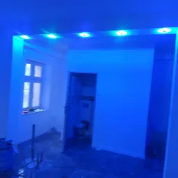 Wnętrze w trakcie remontu, z nową instalacją oświetleniową LED w kolorze niebieskim, widoczne narzędzia i butla gazowa, okno z widokiem na zewnątrz.